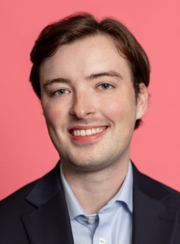 A young man with short, brown hair and a light complexion smiles at the camera. He is wearing a dark blazer over a light blue button-up shirt, with a pink background behind him.